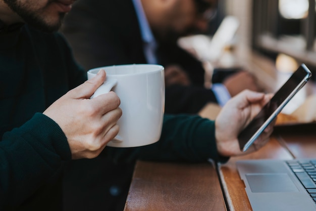 Uma pessoa segurando uma xícara de café. O foco é só na mão. A xícara é branca e lisa, de cerâmica brilhante. A pessoa usa um suéter verder escuro, e segura a xícara com uma mão só. A outra segura um celular apoiada em uma mesa de madeira. Apesar de desfocada, dá pra ver outra pessoa sentada na mesa perto dela, como se eles estivessem em um lugar público a céu aberto, em um clima frio. Todos estão agasalhados e a xícara reforça essa impressão de que 'estão se esquentando'. O tema do blog é Garrafa térmica: amiga ou inimiga do café? 